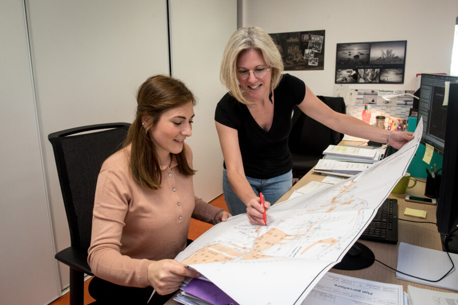 Deux femmes dans un bureau examinent une grande carte. Lune, assise dans un pull rose, écoute attentivement tandis que lautre, vêtue dune chemise noire et brandissant un stylo rouge, pointe avec précision les détails fonciers. Le bureau est encombré de papiers et de fournitures de bureau.
