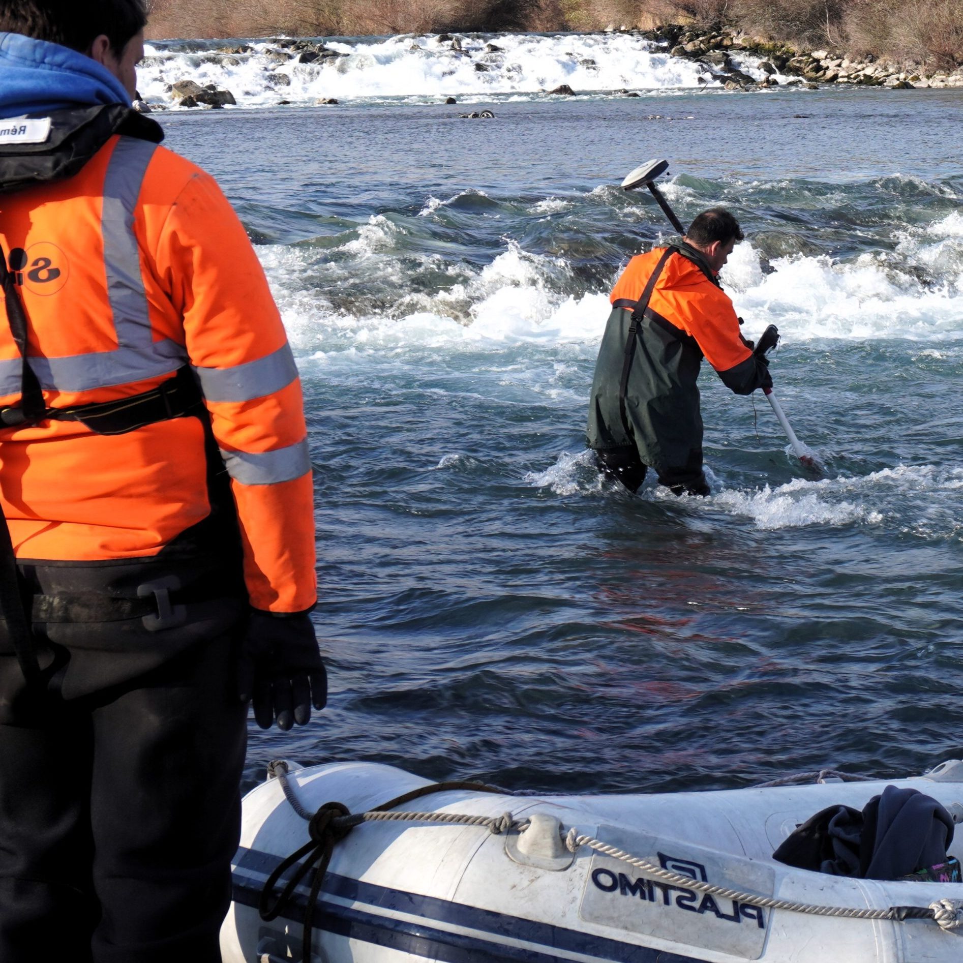 Deux hommes en veste orange effectuent une mesure bathymétrique dune rivière. Lun se tient sur la rive et tient un équipement, tandis que lautre est dans leau avec une perche de mesure. Un radeau se trouve au premier plan et une petite cascade tombe doucement en arrière-plan.