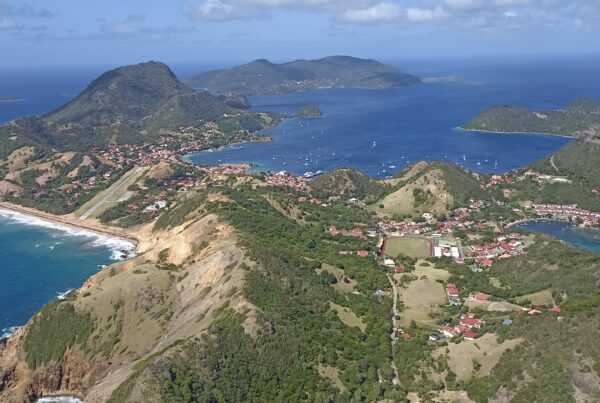 Vue aérienne d'un paysage côtier ponctué de collines verdoyantes, d'un littoral sinueux et de bâtiments aux toits rouges dispersés. Alors que les préparatifs des JO 2024 se déroulent, l'océan bleu entoure plusieurs petites îles au loin, sous un ciel partiellement nuageux.