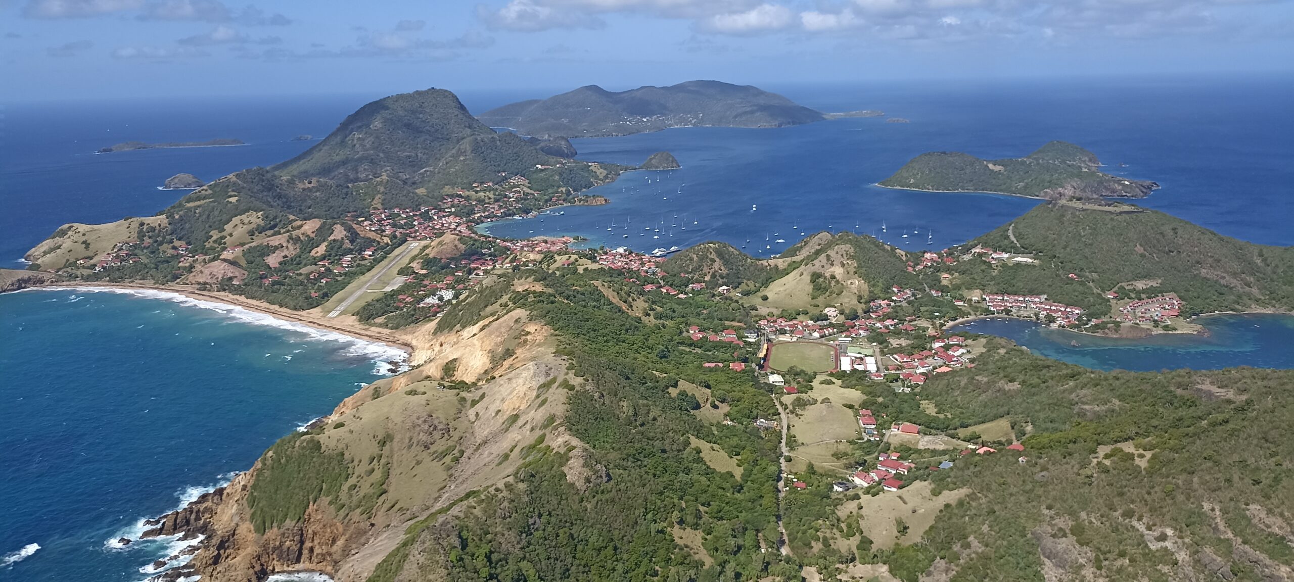 Vue aérienne d'un paysage côtier ponctué de collines verdoyantes, d'un littoral sinueux et de bâtiments aux toits rouges dispersés. Alors que les préparatifs des JO 2024 se déroulent, l'océan bleu entoure plusieurs petites îles au loin, sous un ciel partiellement nuageux.