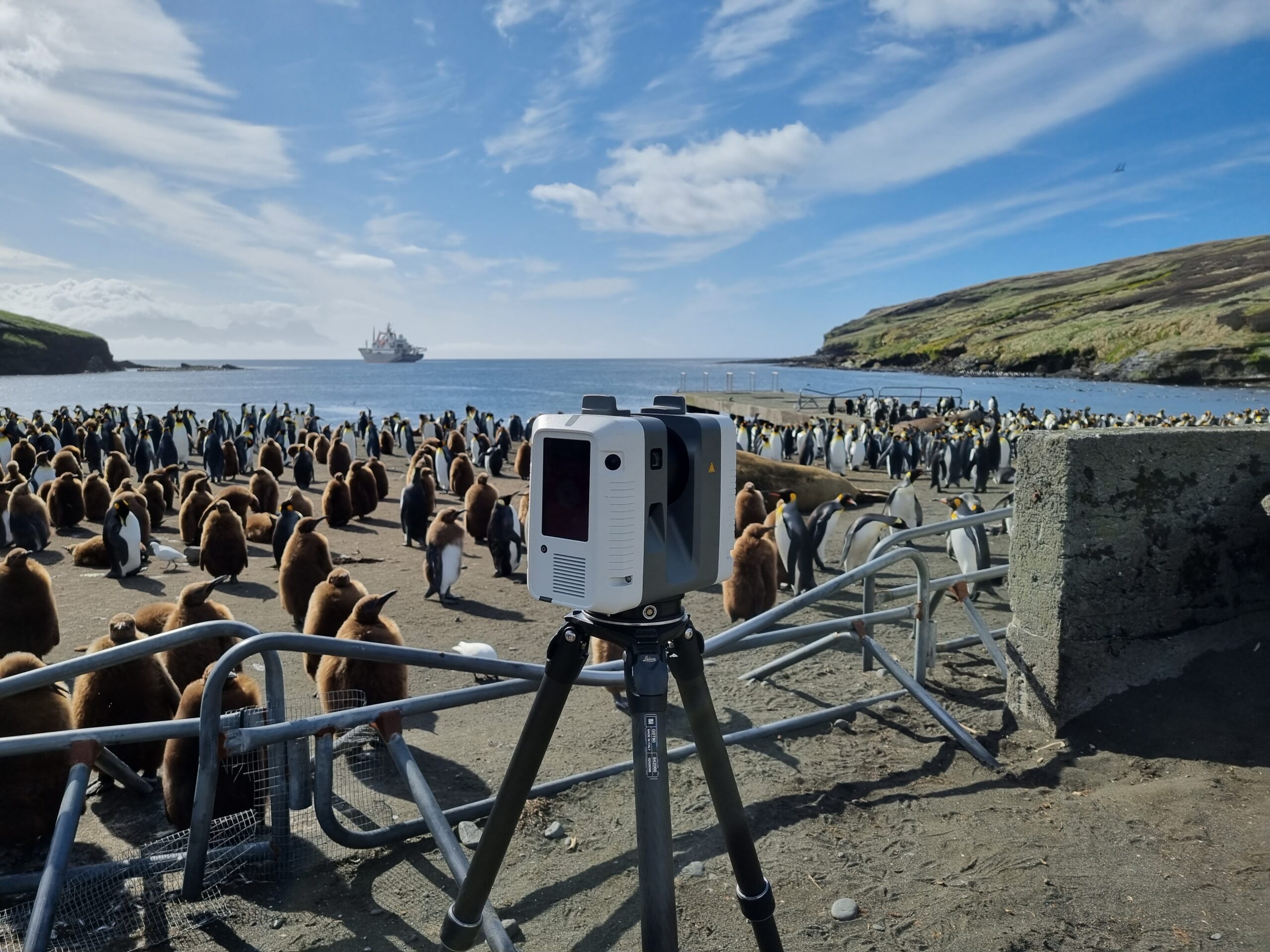 Un scanner 3D sur trépied est installé devant une grande colonie de manchots sur une plage des TAAF : Les Terres australes et antarctiques françaises. Locéan et un navire sont visibles en arrière-plan sous un ciel partiellement nuageux, avec des barrières métalliques entourant la zone du scanner.