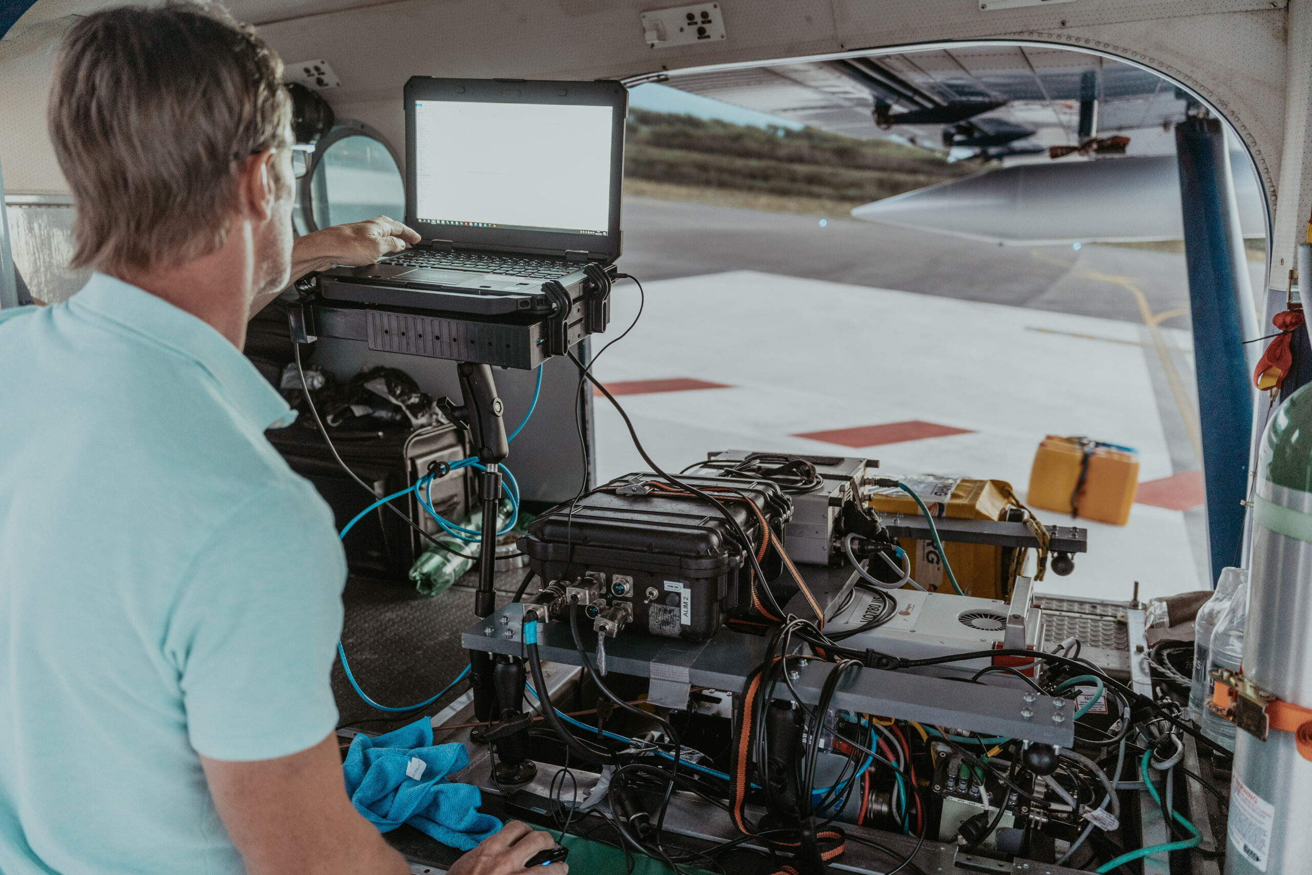 Une personne en chemise bleu clair travaille sur un ordinateur portable relié à divers appareils électroniques à lintérieur dun avion, occupée à un projet dacquisition aérienne. La porte de lavion est ouverte, révélant le tarmac et la piste à lextérieur. Des câbles et des équipements sont éparpillés un peu partout.