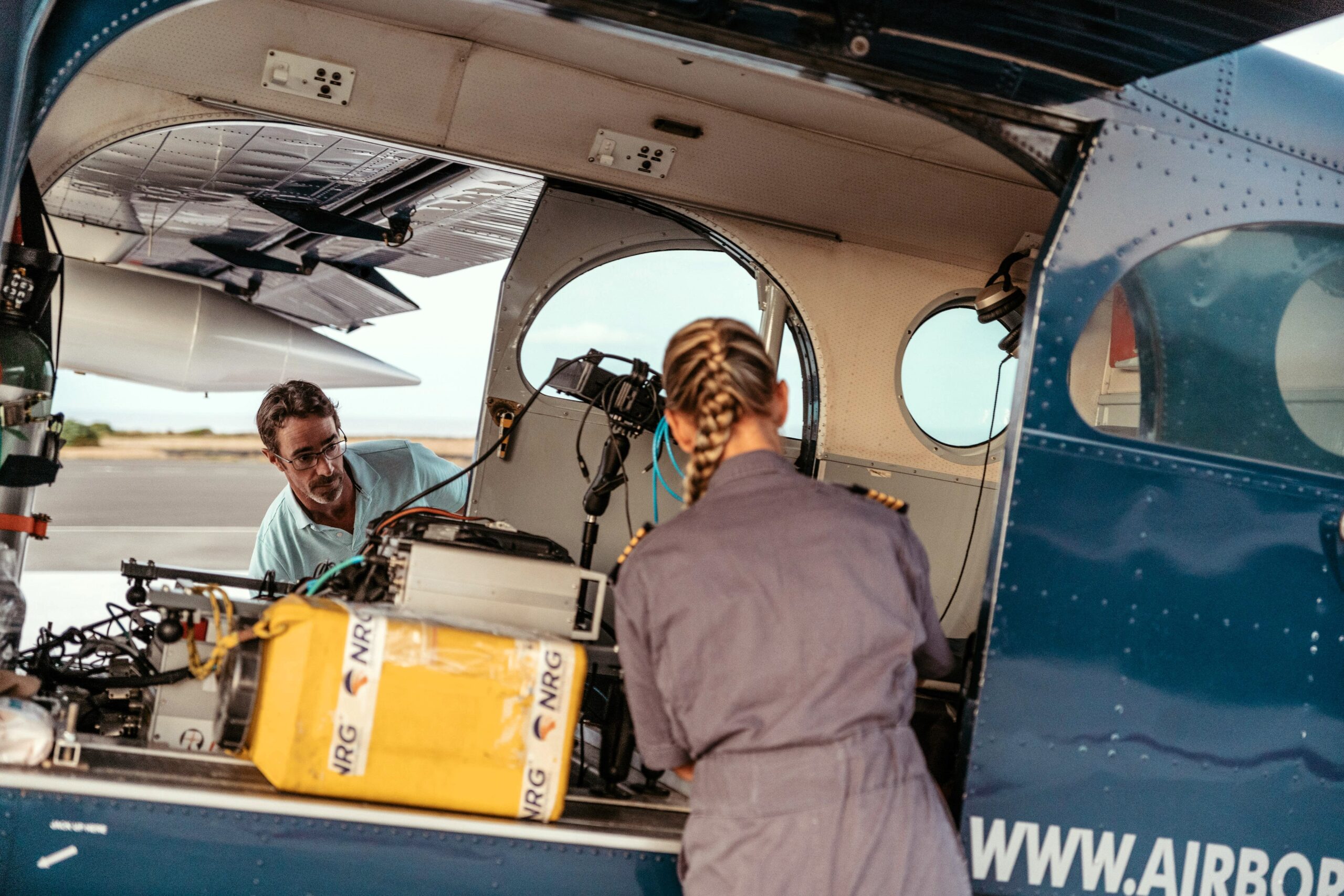 Deux personnes inspectent du matériel à lintérieur de la porte ouverte dun petit avion. La personne au premier plan a les cheveux tressés et porte un uniforme. Divers appareils électroniques dacquisition aérienne, dont un LiDAR photo, ainsi que des fils sont visibles à lintérieur de lavion.