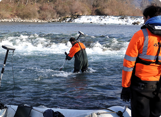 Deux hommes en veste orange effectuent une mesure bathymétrique dune rivière. Lun se tient sur la rive et tient un équipement, tandis que lautre est dans leau avec une perche de mesure. Un radeau se trouve au premier plan et une petite cascade tombe doucement en arrière-plan.