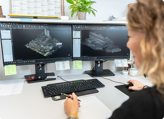 Une personne travaille à un bureau avec deux écrans dordinateur présentant des modèles 3D darchitecture ou de modélisation. Un clavier, une souris et du papier avec des notes écrites sont posés sur le bureau, tandis quune plante et des fournitures de bureau créent une ambiance de fond.