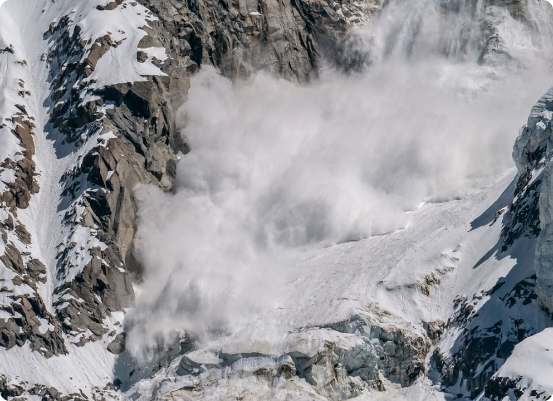 Une avalanche de neige dévale une pente abrupte et rocheuse, créant un nuage de neige et de débris. Le terrain accidenté, semé dembûches, présente des plaques de neige blanche qui contrastent fortement avec les surfaces rocheuses grises.