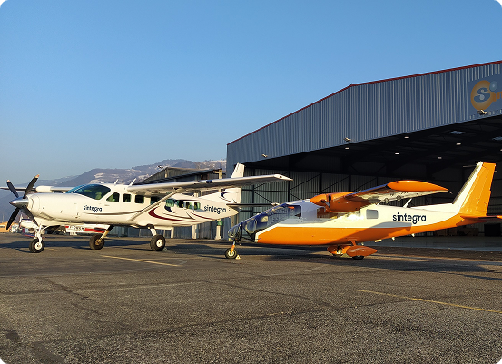 Deux petits avions étiquetés « sintegra » sont stationnés sur le tarmac dun aéroport, devant limpressionnant Hangar de SINTEGRA. Lun des avions est blanc, tandis que lautre arbore un design orange et blanc saisissant. Un ciel clair et des collines lointaines encadrent le fond serein.