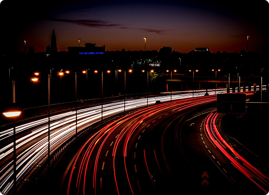Photographie en exposition longue dune autoroute la nuit, capturant les traînées dynamiques de lumières rouges et blanches des véhicules en mouvement. Les lampadaires illuminent les artères de transport sous un horizon urbain sombre se découpant sur le ciel crépusculaire orange et bleu.
