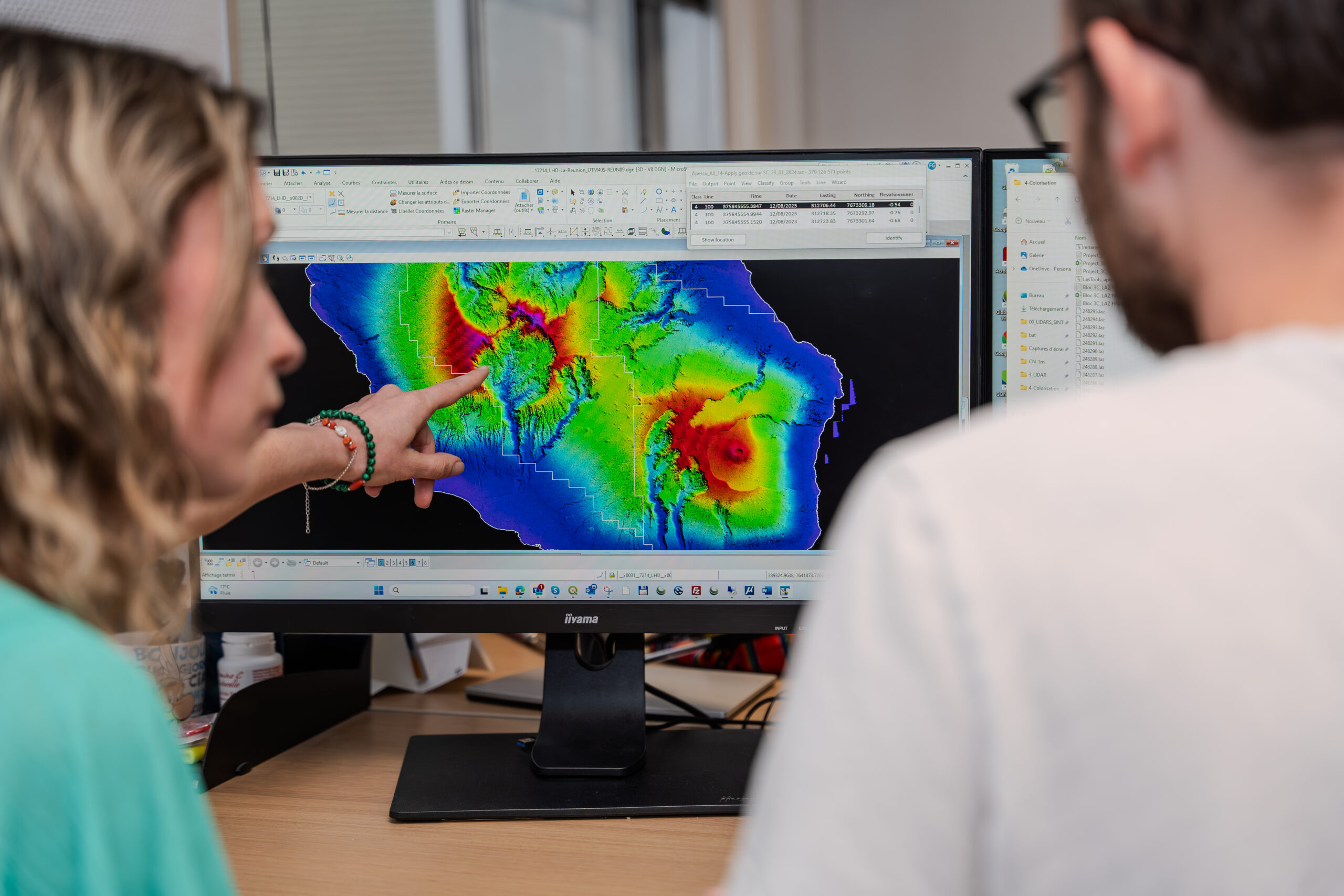Deux personnes sont assises à un bureau et examinent des données topographiques colorées sur un écran. Une personne pointe du doigt des détails spécifiques liés au traitement photo LiDAR. Lespace de travail est rempli darticles de bureau essentiels et déquipements informatiques, créant une atmosphère propice à une analyse détaillée.