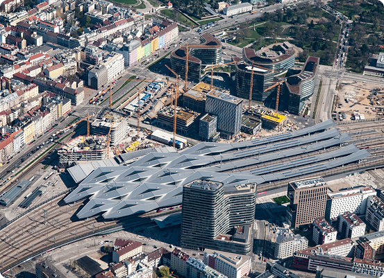 Vue aérienne dune gare moderne avec un toit géométrique caractéristique, entourée de grues, de chantiers de construction et de bâtiments urbains. Les routes et espaces verts à proximité illustrent laménagement stratégique du territoire, mettant en valeur une zone urbaine en cours de développement.