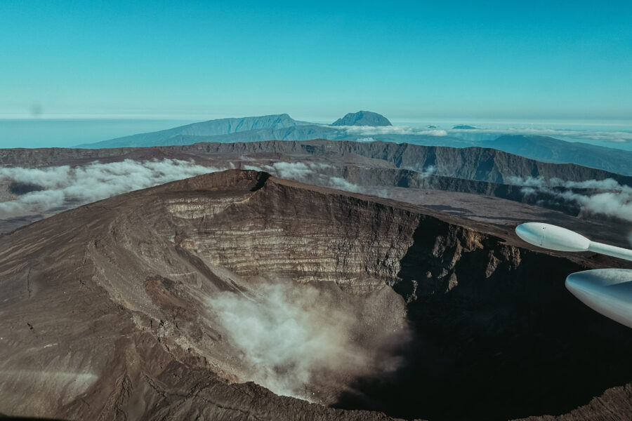 Une vue aérienne d'un grand cratère volcanique rocheux à La Réunion révèle un paysage accidenté. De fins nuages dérivent près du cratère, avec des montagnes au loin sous un ciel bleu clair. Une partie de l'aile d'un avion est visible à droite.