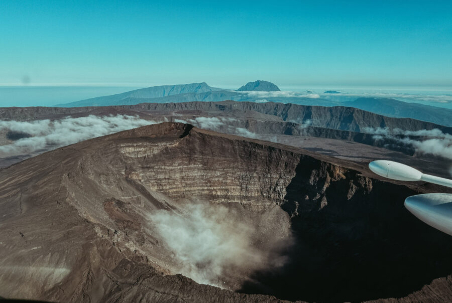 Une vue aérienne d'un grand cratère volcanique rocheux à La Réunion révèle un paysage accidenté. De fins nuages dérivent près du cratère, avec des montagnes au loin sous un ciel bleu clair. Une partie de l'aile d'un avion est visible à droite.