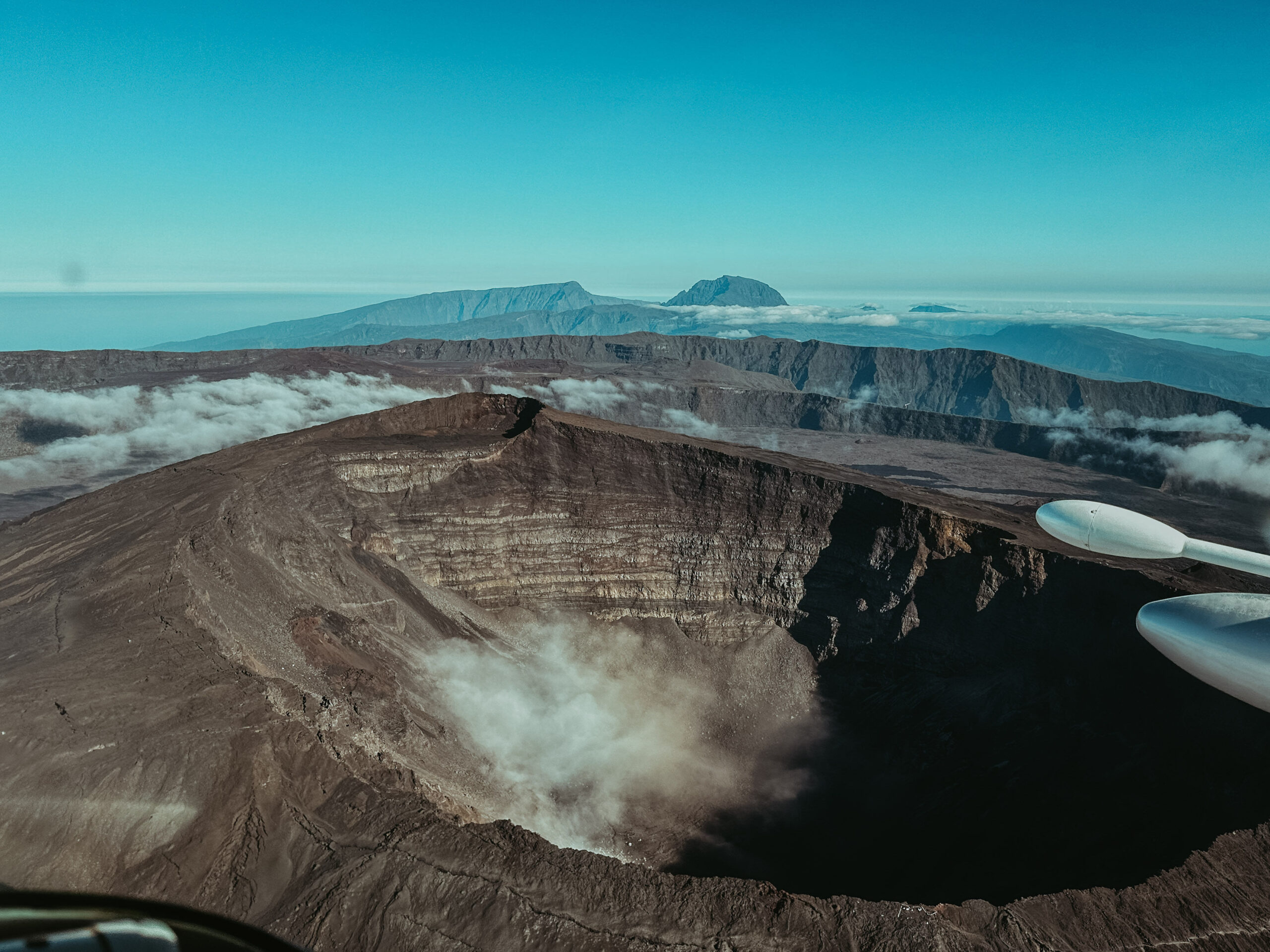 Une vue aérienne d'un grand cratère volcanique rocheux à La Réunion révèle un paysage accidenté. De fins nuages dérivent près du cratère, avec des montagnes au loin sous un ciel bleu clair. Une partie de l'aile d'un avion est visible à droite.