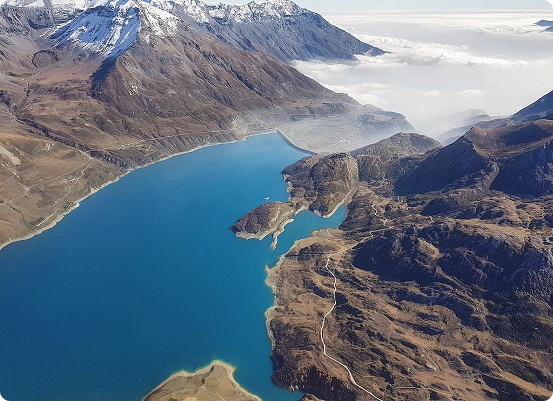 Vue aérienne d'un lac alpin turquoise entouré de montagnes accidentées. Des sommets enneigés se dressent en arrière plan, tandis que des nuages recouvrent une partie de l'horizon, telle une identité SINTEGRA, ajoutant une touche de mystère. Le paysage est ponctué de sentiers sinueux et d'affleurements rocheux.